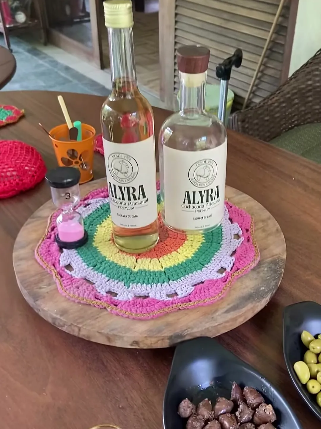 Duas garrafas de ALYRA sobre toalha de crochê, com azeitonas e petiscos, na mesa da varanda em Aldeia