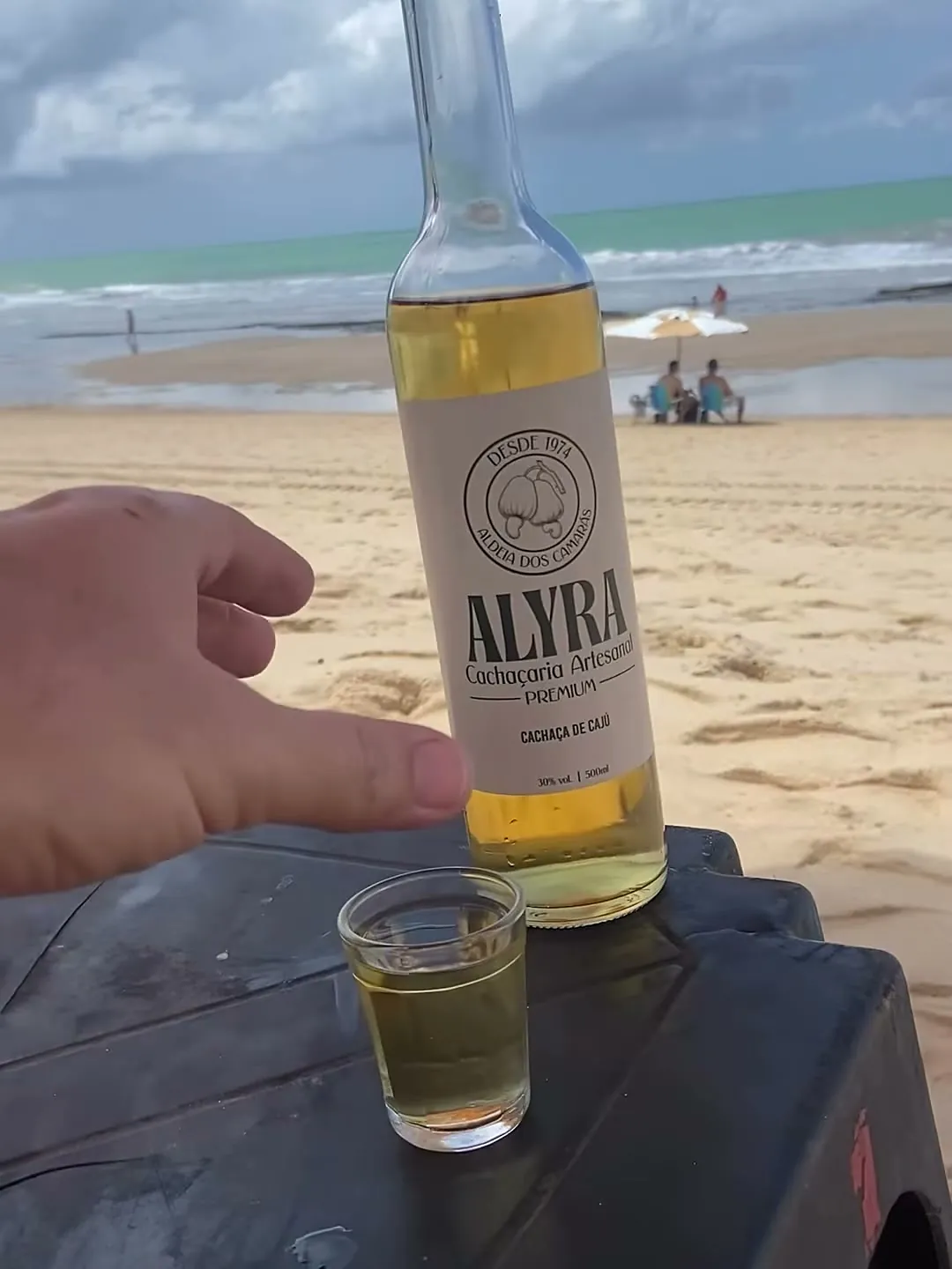 Garrafa e dose de ALYRA numa mesinha na areia da praia
